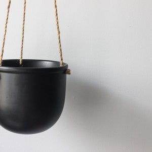 May include: A matte black hanging planter with a rounded bowl shape. The planter features three holes along the upper rim, threaded with natural tan jute rope for suspension. The design is simple and functional, providing a clean look for plants.