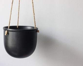 May include: A matte black hanging planter with a rounded bowl shape. The planter features three holes along the upper rim, threaded with natural tan jute rope for suspension. The design is simple and functional, providing a clean look for plants.