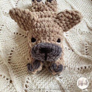 May include: A crocheted plush toy in the shape of a French Bulldog, lying on a white knitted blanket. The toy is brown and gray with a black nose and black eyes.