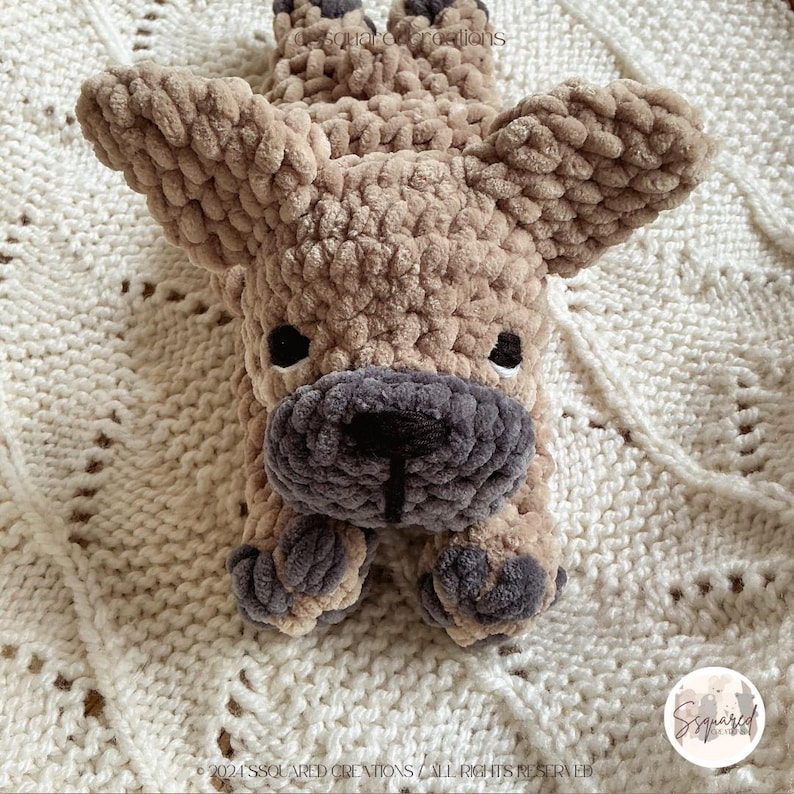 May include: A crocheted plush toy in the shape of a French Bulldog, lying on a white knitted blanket. The toy is brown and gray with a black nose and black eyes.
