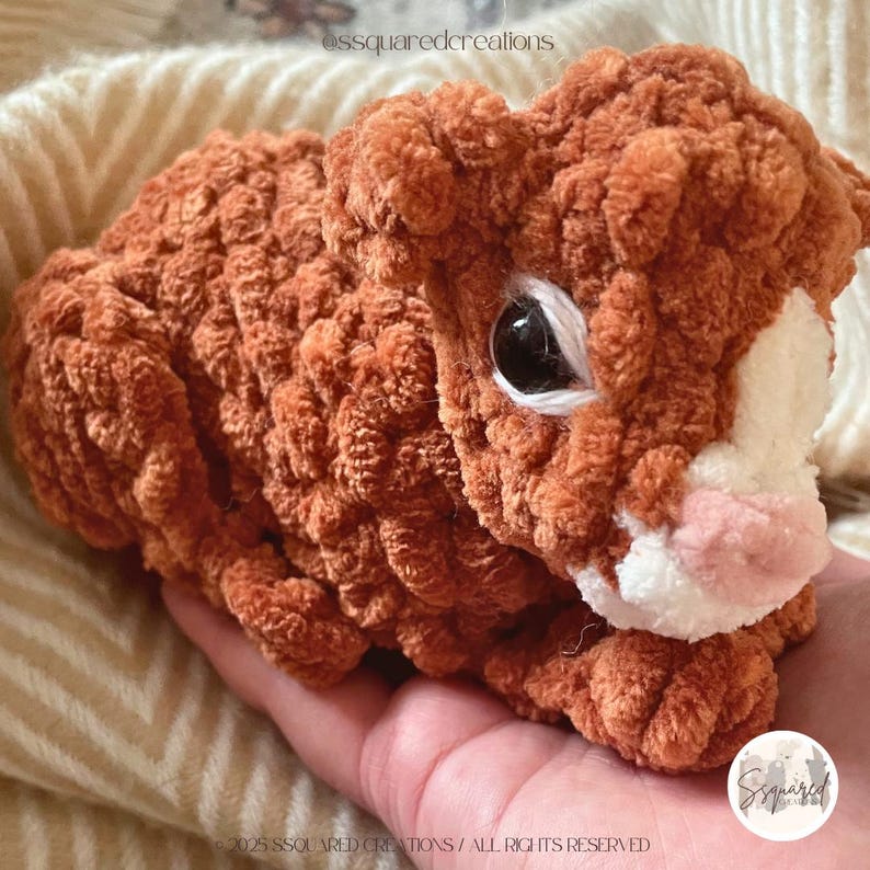 May include: A brown and white crocheted stuffed animal with large eyes. The animal is lying on a hand.