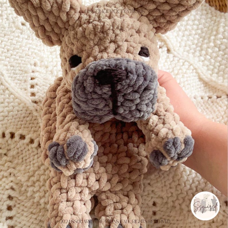 May include: A crocheted plush toy dog in the shape of a French Bulldog. The dog is light brown with a gray face and paws. The dog is being held by a hand.