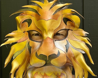 May include: A handcrafted lion mask with a golden yellow and orange color scheme. The mask features intricate details, including a mane, eyes, and a beard. The mask is made of a flexible material and is designed to be worn.