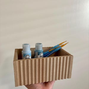 May include: A wooden box with a corrugated exterior contains two small bottles of blue paint and several blue paintbrushes. The bottles have white labels with black text. The paintbrushes have orange bristles.