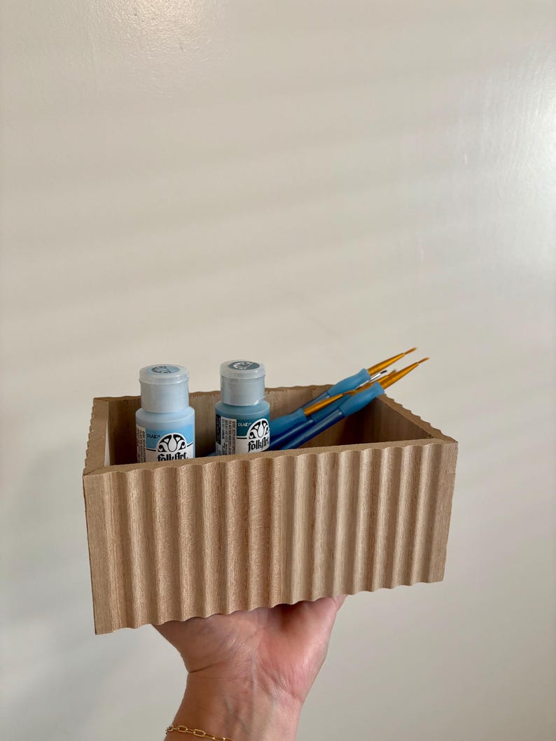 May include: A wooden box with a corrugated exterior contains two small bottles of blue paint and several blue paintbrushes. The bottles have white labels with black text. The paintbrushes have orange bristles.