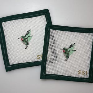 May include: Two square needlepoint coasters with a white background and a green border. Each coaster features a hummingbird design in shades of green, black, and red. The initials "SSI" are stitched in gold in the lower right corner.