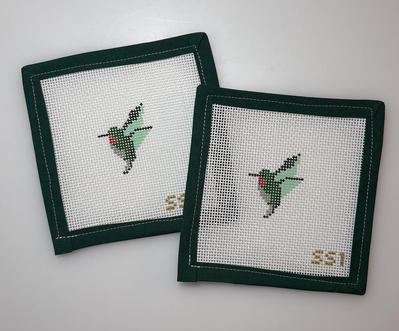 May include: Two square needlepoint coasters with a white background and a green border. Each coaster features a hummingbird design in shades of green, black, and red. The initials "SSI" are stitched in gold in the lower right corner.