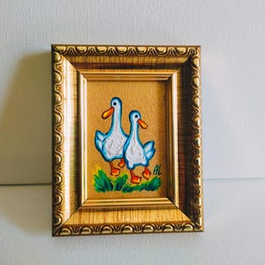 May include: A framed painting of two white ducks with blue beaks and feet walking on a green grassy field. The painting is set against a tan background and is framed in a gold frame.