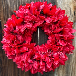 Puede incluir: Una vibrante corona floral roja, densamente llena de pétalos en capas, sobre un fondo de madera rústica. La corona es circular, con una abertura central, y presenta el texto "Old Road Wreaths".