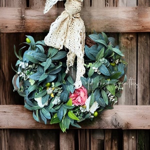 Könnte beinhalten: Ein handgefertigter Kranz mit künstlichen Olivenzweigen, grünen Blättern und einer großen rosa Rose. Akzentuiert mit weißen Blüten und grünen Beeren. Ein cremefarbener Spitzenbogen ziert den Kranz. Der Text "old road wreaths" ist sichtbar.