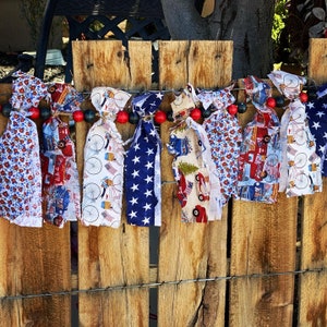 May include: A patriotic fabric garland with red, white, and blue patterns, including stars, bicycles, and floral prints. The garland is strung with red and black wooden beads and hangs against a rustic wooden fence.