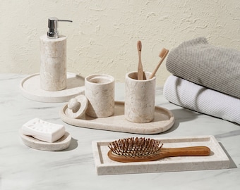 May include: A set of bathroom accessories in a light beige marble finish. The set includes a soap dispenser, a toothbrush holder, a cotton swab jar, a soap dish, and a hairbrush tray. The accessories are arranged on a white countertop.
