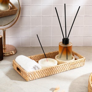 May include: A woven wicker tray with a white towel, a small white bowl, a glass bottle with black diffuser sticks, and a wooden incense stick.