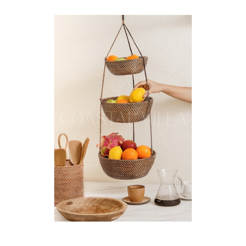 May include: A three-tiered hanging fruit basket made of woven rattan with a dark brown finish. The basket is filled with fresh fruit, including oranges, lemons, and a dragon fruit. The basket is hanging from the ceiling with brown cords.
