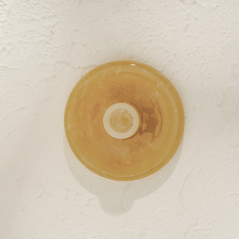 May include: A round, yellow glass candle holder with a white center. The candle holder is on a white textured surface.