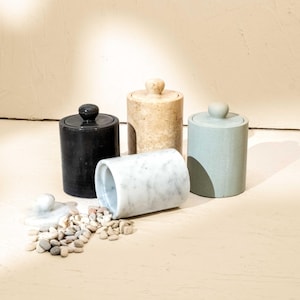 May include: Four cylindrical stone canisters with matching lids. One is solid black, one is beige with a mottled texture, one is sage green, and one is white marble lying on its side. A small pile of smooth, light-coloured pebbles sits nearby.
