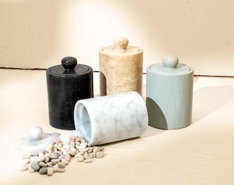 May include: Four cylindrical stone canisters with matching lids. One is black, one is beige, one is light green, and one is white with gray veining, lying on its side. A small pile of smooth, light-colored pebbles sits near the white canister.