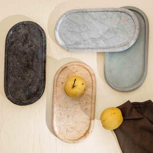 Puede incluir: Cuatro bandejas de forma ovalada en diversos materiales y colores, incluyendo una piedra gris oscuro, mármol blanco con vetas grises, travertino beige claro y una bandeja de hormigón gris, con dos peras amarillas colocadas sobre ellas.
