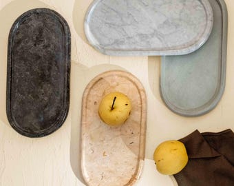 May include: Four oval-shaped trays in varying colors and textures, including a dark charcoal stone, a white marble, a light beige stone, and a solid grey tray. Two yellow pears are placed on the beige and grey trays respectively.