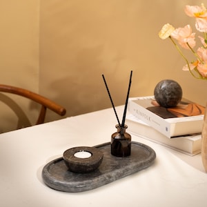 Puede incluir: Una bandeja ovalada de piedra gris oscuro que contiene un portavelas circular a juego con una vela de té blanca y un pequeño frasco difusor de vidrio marrón con dos varillas negras. El conjunto se sitúa sobre una superficie blanca junto a libros.