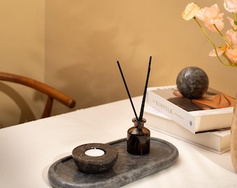 May include: An oval dark gray stone tray holding a small round stone candle holder with a white tea light and a dark brown glass bottle containing black reed diffuser sticks. The tray is positioned on a white surface near stacked books.