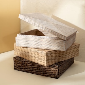May include: A stack of three rectangular woven storage boxes with lids. The top box is white, the middle box is a natural light tan, and the bottom box is dark brown. Each box features a tight, uniform weave pattern and a flat, square lid.