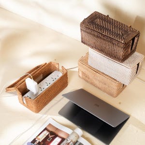 Könnte beinhalten: Eine Sammlung rechteckiger Kabelboxen aus geflochtenem Rattan in Dunkelbraun, hellem Beige und Naturfarben. Eine geöffnete Box enthält eine weiße Steckdosenleiste mit eingestecktem Ladegerät. Ein geschlossener Laptop und ein Magazin liegen daneben.