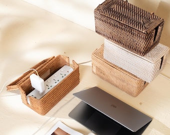 May include: A collection of rectangular woven rattan cable management boxes in dark brown, light beige, and natural tan. One open box contains a white power strip with a plugged-in charger. A closed laptop and a magazine are placed nearby.