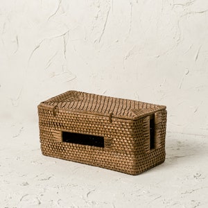 May include: A woven wicker tissue box cover with a rectangular shape and a cut-out opening on the front. The cover is a light brown colour.