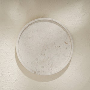 May include: A circular tray crafted from light beige stone with a natural mottled texture and visible veining. The tray features a raised rim and a smooth, flat surface, showcasing the organic variations and earthy tones inherent in the stone material.
