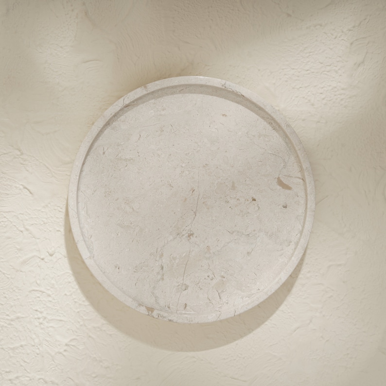 May include: A circular tray crafted from light beige stone with a natural mottled texture and visible veining. The tray features a raised rim and a smooth, flat surface, showcasing the organic variations and earthy tones inherent in the stone material.