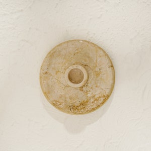 May include: A round, beige marble candle holder with a small, circular opening in the center.