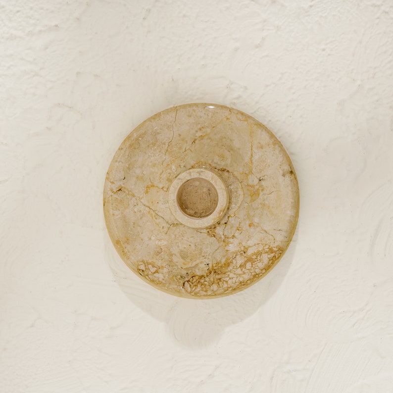 May include: A round, beige marble candle holder with a small, circular opening in the center.