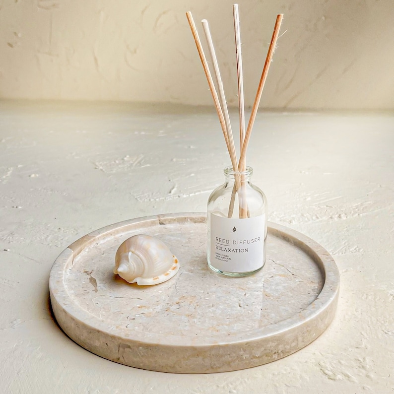 May include: A clear glass reed diffuser bottle with four wooden reeds and a white label reading REED DIFFUSER RELAXATION, placed on a round beige stone tray next to a small, smooth, cream-colored seashell with subtle orange-brown spots.