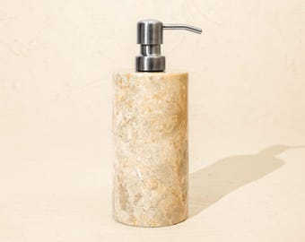 May include: A cylindrical soap dispenser crafted from beige and tan mottled stone with a smooth, polished finish. The top features a brushed silver-toned metal pump mechanism with a horizontal nozzle extending to the right side of the dispenser.
