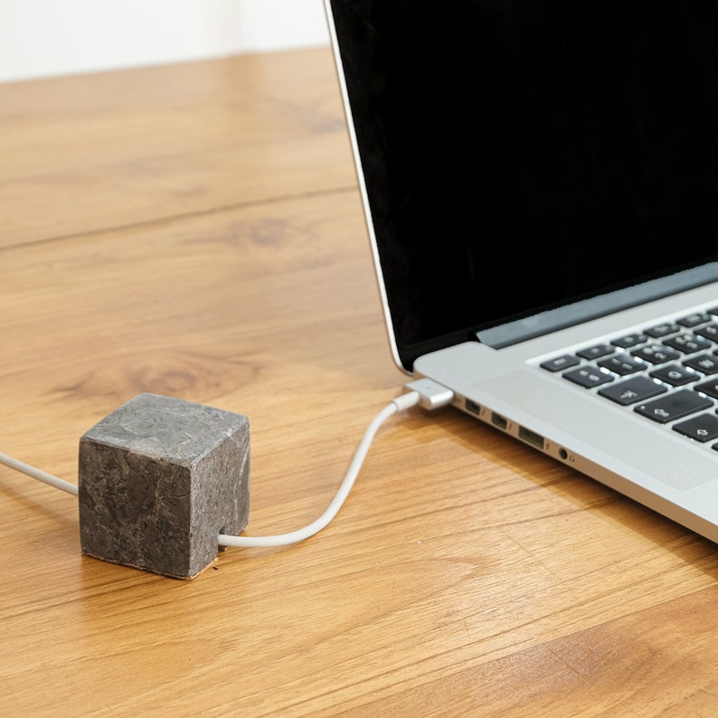 May include: A gray stone cable organizer on a wooden surface with a white cable running to a laptop computer.