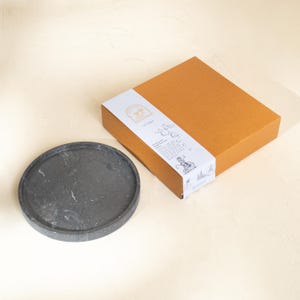 May include: A circular dark gray stone tray with subtle white veining and a smooth finish, positioned next to a square tan cardboard gift box wrapped in a white paper sleeve featuring black line art illustrations and printed text.