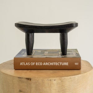 Könnte beinhalten: Ein schwarzer Holzhocker mit vier Beinen, der auf einem Buch mit dem Titel "Atlas of Eco Architecture" steht.