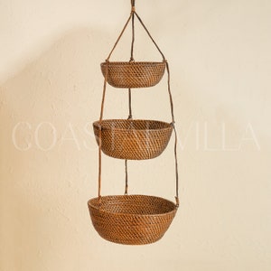 May include: A three-tiered hanging wicker basket set with a natural brown finish. The baskets are suspended from a ceiling or wall by a set of cords.