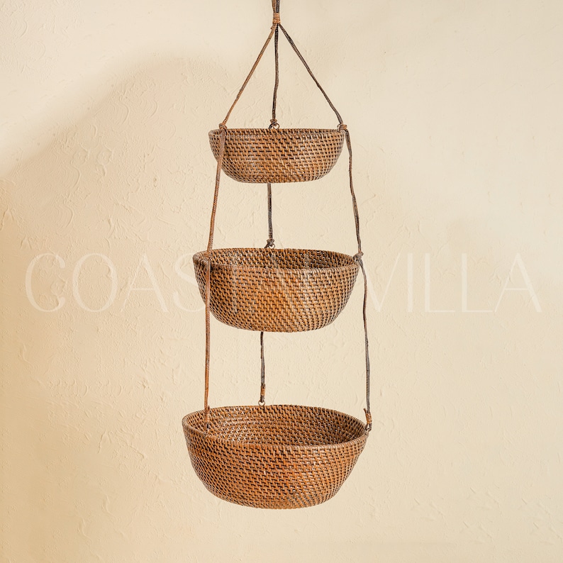 May include: A three-tiered hanging wicker basket set with a natural brown finish. The baskets are suspended from a ceiling or wall by a set of cords.
