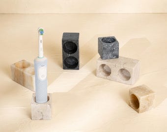 May include: A light blue electric toothbrush stands in a beige stone holder. Nearby are several other stone holders in beige, white, and black, featuring various cylindrical cutouts for storage, including rectangular and square block shapes.