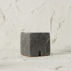 May include: A small, square, black marble pedestal with a cork base. The pedestal is sitting on a white textured surface.