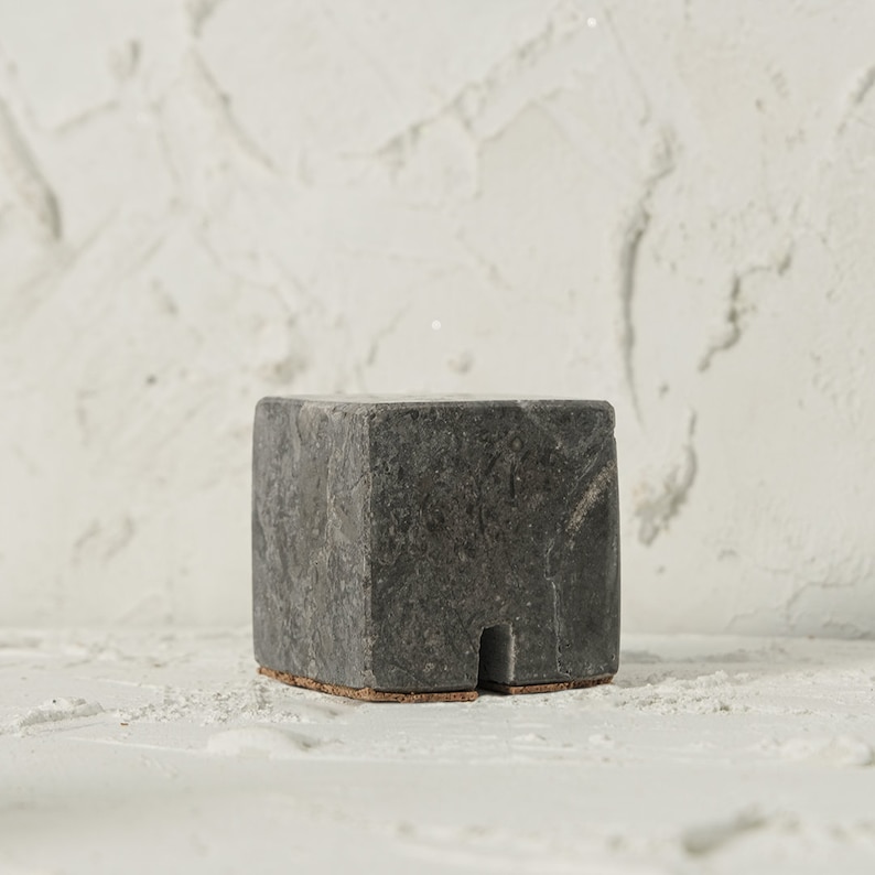 May include: A small, square, black marble pedestal with a cork base. The pedestal is sitting on a white textured surface.
