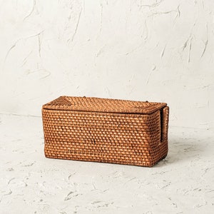 May include: A rectangular woven wicker storage basket with a lid. The basket is made of natural brown rattan and has a woven pattern.