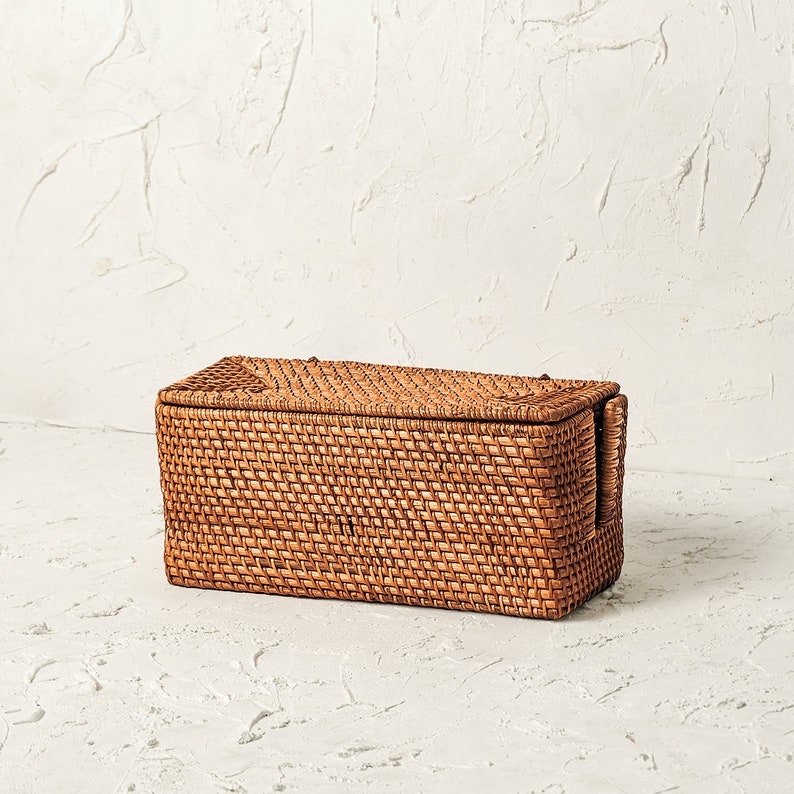 May include: A rectangular woven wicker storage basket with a lid. The basket is made of natural brown rattan and has a woven pattern.