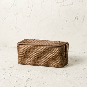 May include: A rectangular woven storage basket with a lid and handles. The basket is made of natural brown wicker and has a woven pattern.