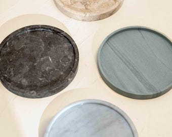 Puede incluir: Cuatro bandejas circulares de piedra con bordes elevados. Una bandeja es beige con textura marmolada, una es gris carbón oscuro con vetas blancas, una es verde salvia apagado con patrones lineales sutiles y una es gris pálido con vetas blancas.