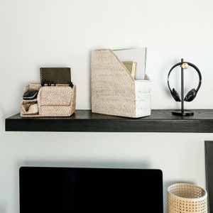 May include: Two white wicker baskets, one with a black mouse and a black phone, the other with a stack of papers, on a black shelf. A black computer monitor and a black and gold headphone stand are on the shelf below.