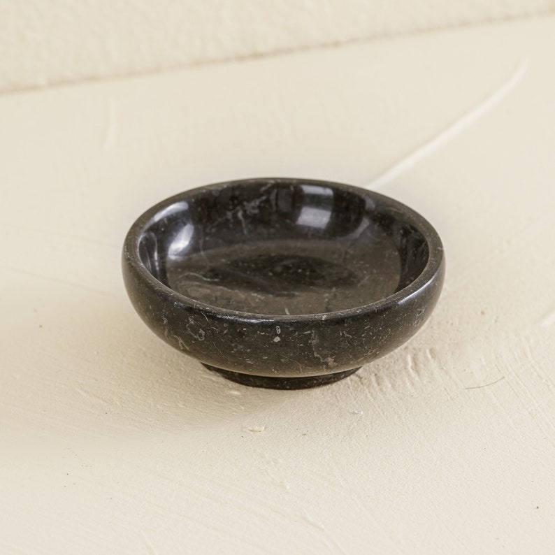 K&ouml;nnte beinhalten: Eine kleine, runde schwarze Marmorschale mit einer glatten, polierten Oberfl&auml;che. Die Schale ist perfekt f&uuml;r Schmuck, Schl&uuml;ssel oder andere kleine Gegenst&auml;nde.