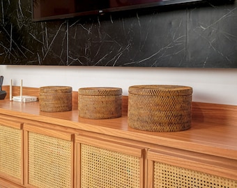 May include: Three brown wicker baskets with lids sit on a wooden shelf with woven cane doors. The baskets are round and have a natural woven pattern.